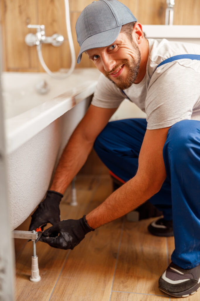 smiling plumber in work gloves tightening a pipe fitting beside a bathtub